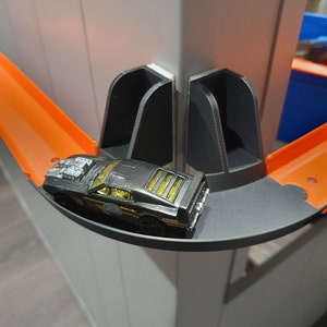 May include: A black toy car with gold accents is driving on a black and orange track. The track is attached to a white wall with black plastic supports.