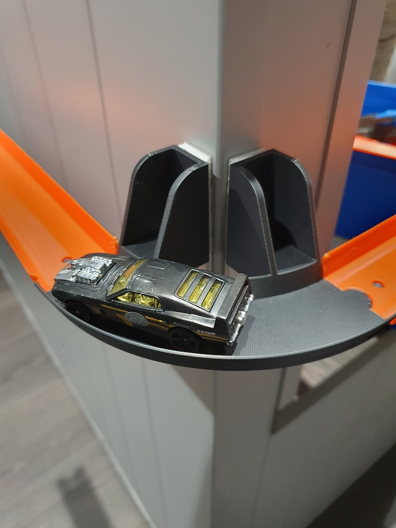 May include: A black toy car with gold accents is driving on a black and orange track. The track is attached to a white wall with black plastic supports.