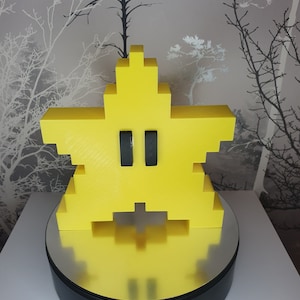 Puede incluir: Una figurita amarilla en forma de estrella con ojos negros, que se asemeja a un personaje de videojuego, sobre una base giratoria negra.