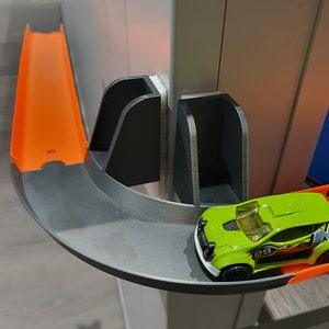 May include: A green toy car with a number 29 on the side is driving on a black and orange plastic track. The track is attached to a white wall with a black plastic bracket.