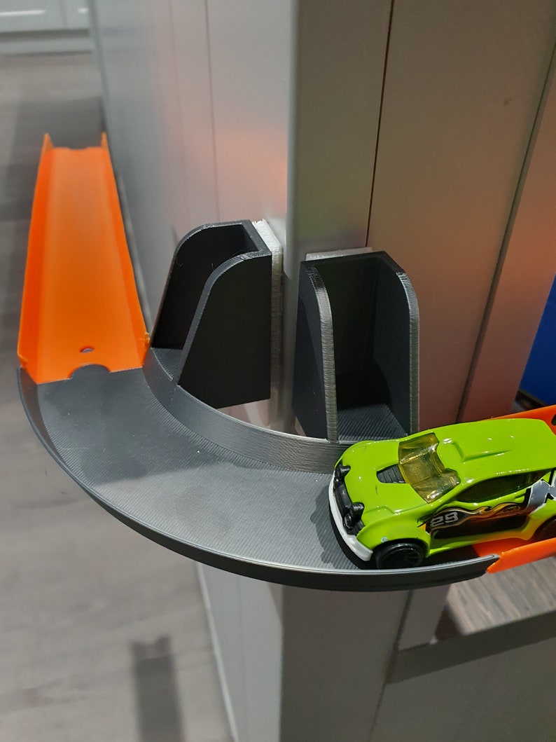 May include: A green toy car with a number 29 on the side is driving on a black and orange plastic track. The track is attached to a white wall with a black plastic bracket.