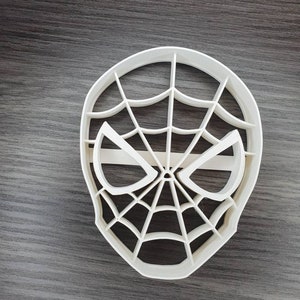 May include: White plastic cookie cutter shaped like a Spiderman mask with a web design.