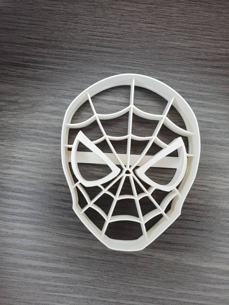 May include: White plastic cookie cutter shaped like a Spiderman mask with a web design.