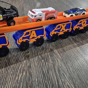 May include: A toy car carrier with a blue and orange design. The carrier is loaded with toy cars, including a police car, a fire truck, and a blue car. The carrier has the letters J, A, X, O, and N on the back.