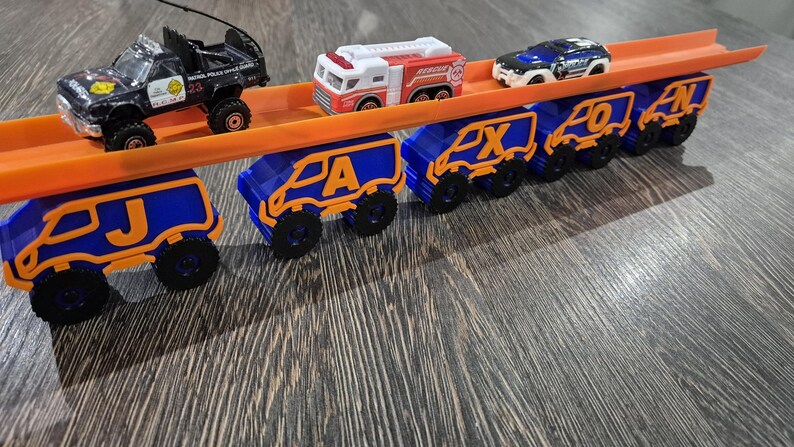 May include: A toy car carrier with a blue and orange design. The carrier is loaded with toy cars, including a police car, a fire truck, and a blue car. The carrier has the letters J, A, X, O, and N on the back.
