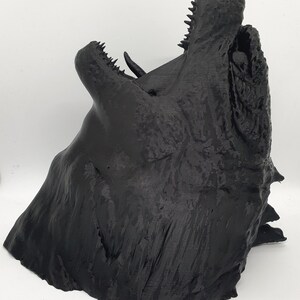 May include: A 3D printed black dragon head sculpture with an open mouth and sharp teeth. The sculpture is detailed and textured, with a rough, layered surface.