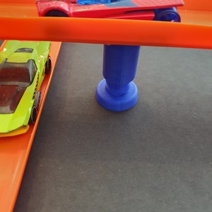 Puede incluir: Un coche de juguete rojo y negro en una pista naranja con un poste de apoyo azul. Un coche de juguete verde est&aacute; en la pista debajo del coche rojo.