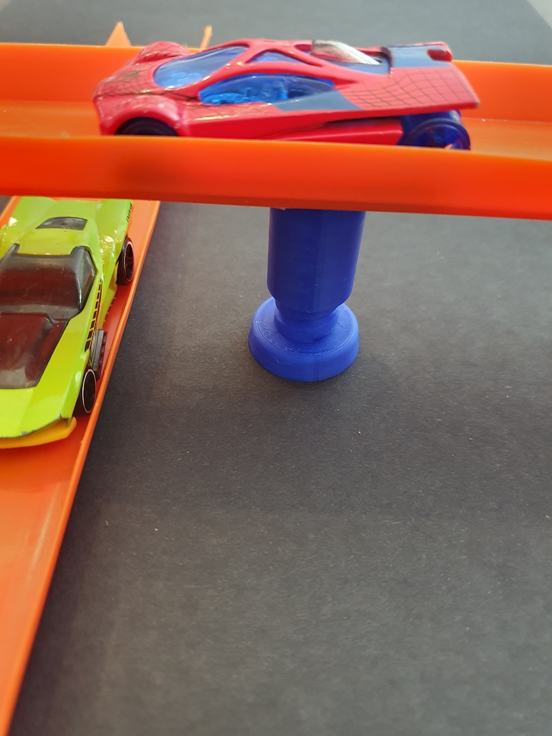 Puede incluir: Un coche de juguete rojo y negro en una pista naranja con un poste de apoyo azul. Un coche de juguete verde est&aacute; en la pista debajo del coche rojo.