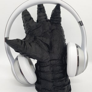 May include: White over-ear headphones with a silver accent on the earcups, resting on a black, textured hand-shaped stand.