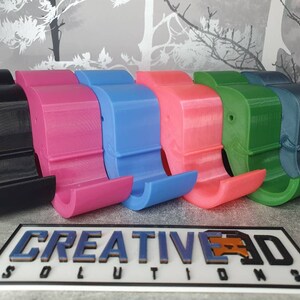 K&ouml;nnte beinhalten: Eine Reihe farbenfroher, 3D-gedruckter Telefonst&auml;nder in Schwarz, Pink, Blau, Koralle, Gr&uuml;n und T&uuml;rkis. Die St&auml;nder haben ein gebogenes Design mit einem Schlitz zur Aufnahme eines Telefons. Das Logo "CREATIVE 3D SOLUTIONS" ist unten zu sehen.