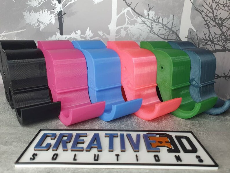 K&ouml;nnte beinhalten: Eine Reihe farbenfroher, 3D-gedruckter Telefonst&auml;nder in Schwarz, Pink, Blau, Koralle, Gr&uuml;n und T&uuml;rkis. Die St&auml;nder haben ein gebogenes Design mit einem Schlitz zur Aufnahme eines Telefons. Das Logo "CREATIVE 3D SOLUTIONS" ist unten zu sehen.