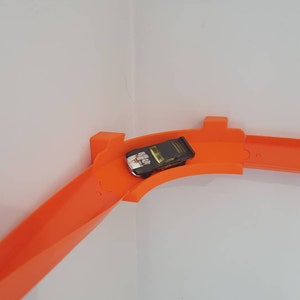 May include: A black toy car on an orange plastic track set. The track is curved and goes around a corner.