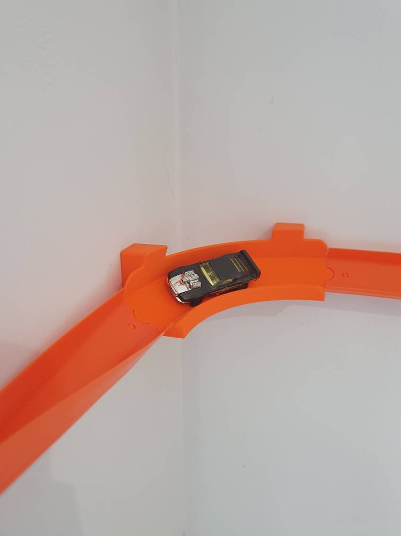 May include: A black toy car on an orange plastic track set. The track is curved and goes around a corner.