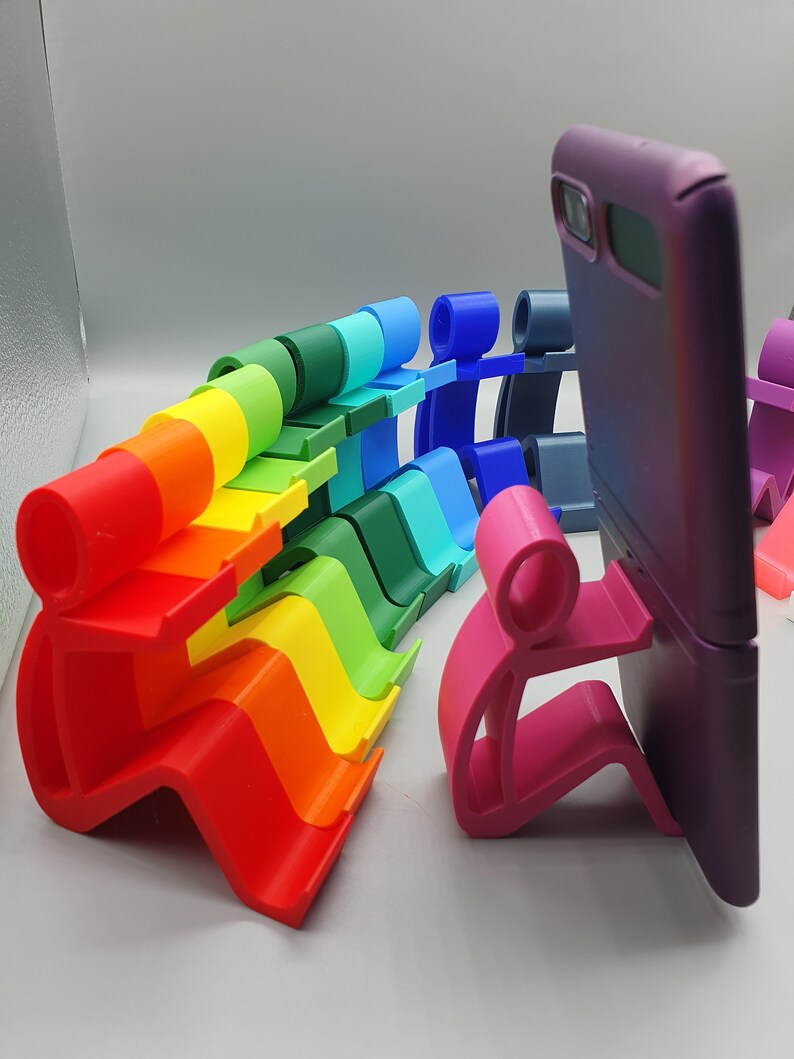 May include: A rainbow of colorful phone stands made of plastic, shaped like a person holding a phone. The stands are arranged in a row, with the pink stand holding a purple phone.