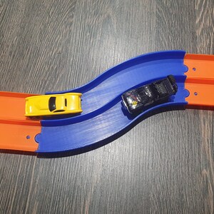 May include: A blue and orange toy race track with two toy cars. One car is yellow and the other is black.