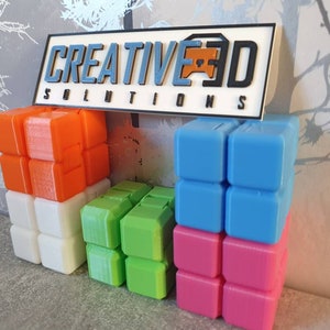 Op de afbeelding: Een verzameling kleurrijke 3D-geprinte blokken gestapeld in verschillende configuraties. De blokken zijn oranje, wit, groen, blauw en roze. Een bord boven de blokken luidt "CREATIVE 3D SOLUTIONS".