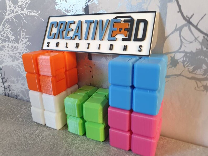 Op de afbeelding: Een verzameling kleurrijke 3D-geprinte blokken gestapeld in verschillende configuraties. De blokken zijn oranje, wit, groen, blauw en roze. Een bord boven de blokken luidt "CREATIVE 3D SOLUTIONS".