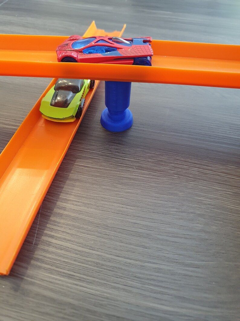 Puede incluir: Dos coches de juguete en un set de pista naranja. El coche rojo est&aacute; en la pista superior y el coche verde est&aacute; en la pista inferior. La pista est&aacute; soportada por un soporte de pl&aacute;stico azul.
