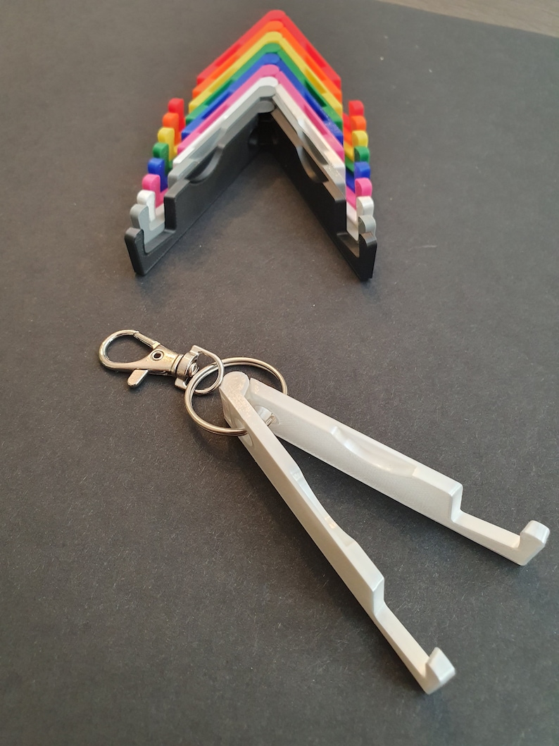 Puede incluir: Un soporte para tel&eacute;fono de metal blanco con un llavero. El soporte est&aacute; plegado y tiene un acabado plateado. El soporte est&aacute; dise&ntilde;ado para sostener un tel&eacute;fono.