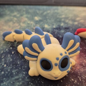 May include: A white and blue 3D printed axolotl figurine with black eyes and blue gills. The figurine is sitting on a blue and white galaxy patterned surface.