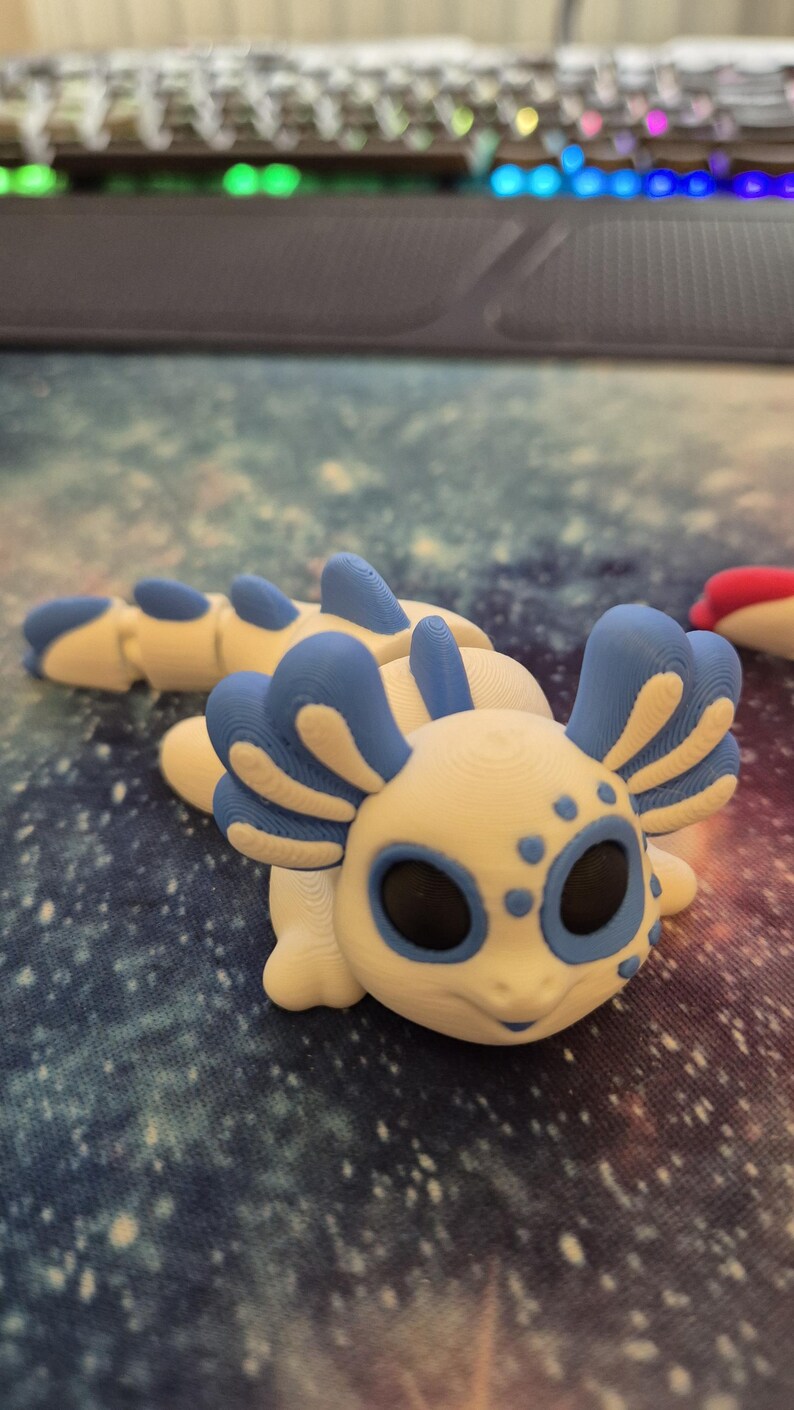 May include: A white and blue 3D printed axolotl figurine with black eyes and blue gills. The figurine is sitting on a blue and white galaxy patterned surface.
