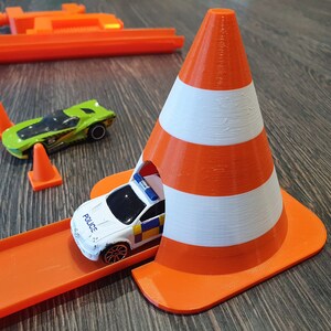 May include: A white and orange toy traffic cone with a toy police car driving through the cone. The car is on an orange track.