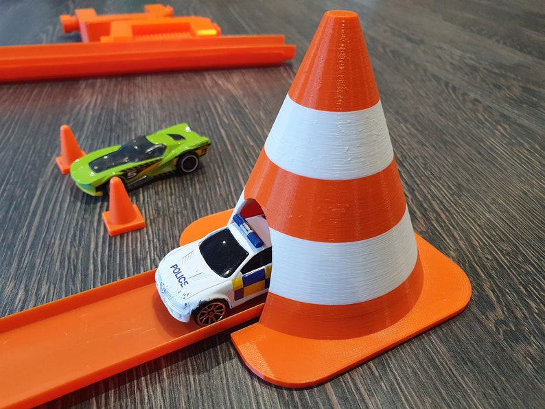 May include: A white and orange toy traffic cone with a toy police car driving through the cone. The car is on an orange track.
