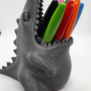 May include: A gray 3D printed dinosaur-shaped pencil holder with a toothy open mouth holding a variety of colored markers, including black, green, red, orange, and blue.