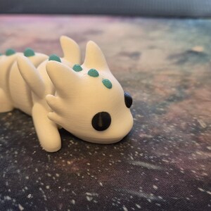 May include: White 3D printed dragon figurine with teal accents and black eyes. The dragon is lying on its belly with its head turned to the side.