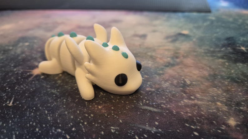 May include: White 3D printed dragon figurine with teal accents and black eyes. The dragon is lying on its belly with its head turned to the side.