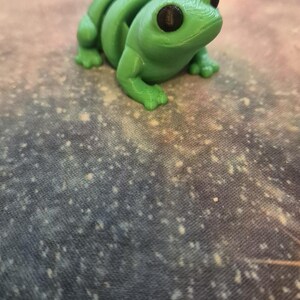 FroggieBum™ Tiny Frog Fidget Keyring: "A small, hand-painted green frog keychain with a round, squishy backside. The frog has a cute, chubby design with tiny legs, sitting on a flat surface. Its smooth, colorful body and charmingly