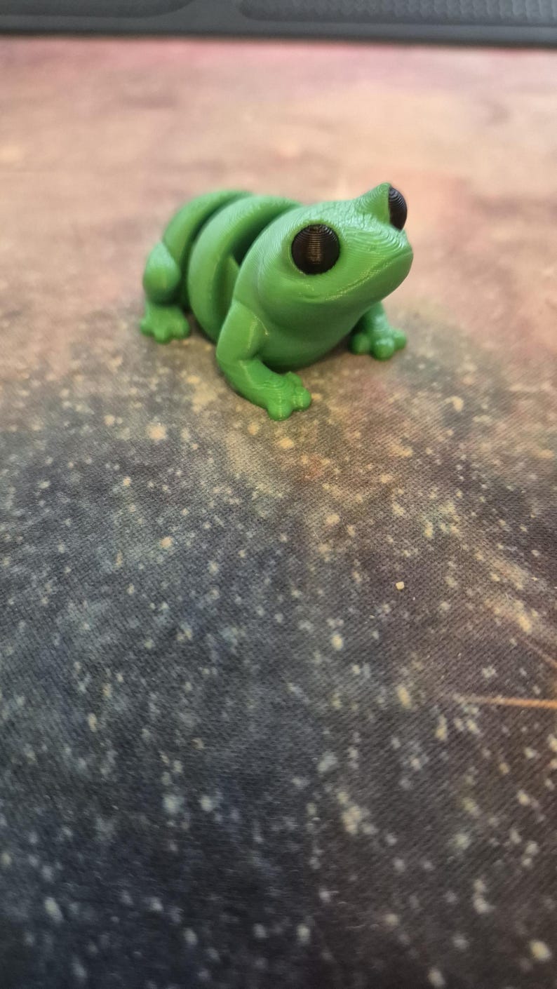 FroggieBum™ Tiny Frog Fidget Keyring: "A small, hand-painted green frog keychain with a round, squishy backside. The frog has a cute, chubby design with tiny legs, sitting on a flat surface. Its smooth, colorful body and charmingly
