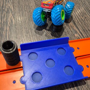 May include: A blue plastic track piece with six holes for a toy monster truck. The track piece is connected to an orange track piece. A red, yellow, and black monster truck with blue wheels is on a separate track piece.