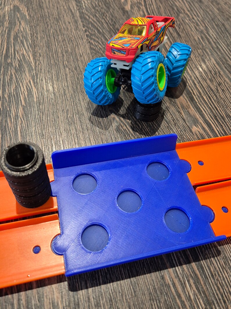 May include: A blue plastic track piece with six holes for a toy monster truck. The track piece is connected to an orange track piece. A red, yellow, and black monster truck with blue wheels is on a separate track piece.