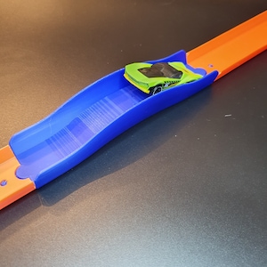 Op de afbeelding: Een blauw plastic baanstuk voor een speelgoedautobaan. Het baanstuk is gebogen en heeft een verhoogd gedeelte in het midden. Een groene speelgoedauto staat op het baanstuk.