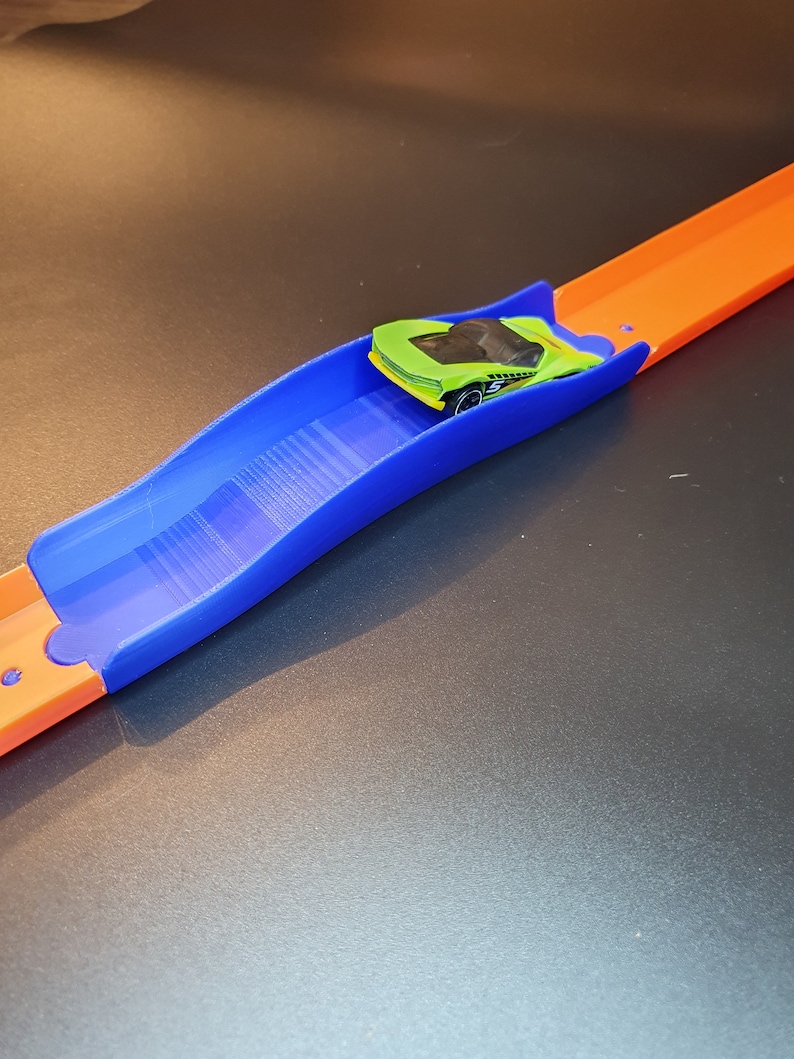 Op de afbeelding: Een blauw plastic baanstuk voor een speelgoedautobaan. Het baanstuk is gebogen en heeft een verhoogd gedeelte in het midden. Een groene speelgoedauto staat op het baanstuk.