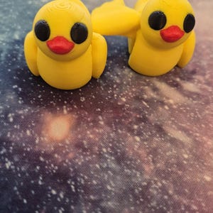 May include: Two yellow rubber duck figurines with black eyes and red beaks. The ducks are facing forward and have a simple design.