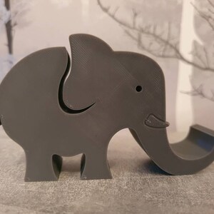 K&ouml;nnte beinhalten: Ein grauer, elefantenf&ouml;rmiger Telefonhalter. Der Elefant hat einen gebogenen R&uuml;ssel, der als Telefonablage dient. Der K&ouml;rper hat einen Schlitz, um das Telefon aufrecht zu halten. Das Design ist schlicht und modern.