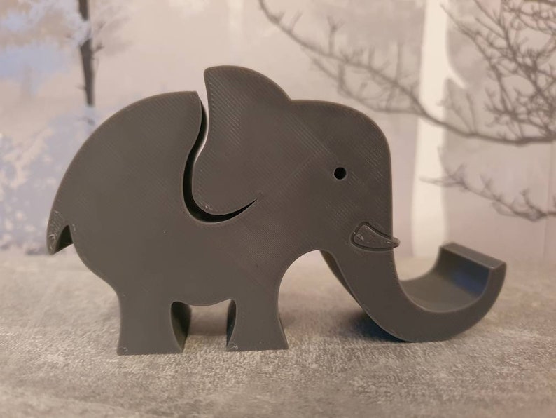 K&ouml;nnte beinhalten: Ein grauer, elefantenf&ouml;rmiger Telefonhalter. Der Elefant hat einen gebogenen R&uuml;ssel, der als Telefonablage dient. Der K&ouml;rper hat einen Schlitz, um das Telefon aufrecht zu halten. Das Design ist schlicht und modern.