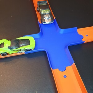 May include: A green and black toy car on an orange track with a blue intersection. A black toy car is on the orange track above the intersection.