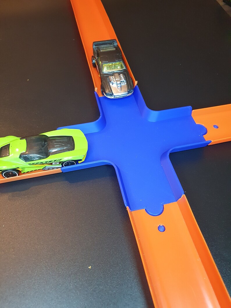 May include: A green and black toy car on an orange track with a blue intersection. A black toy car is on the orange track above the intersection.