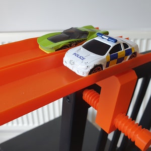 May include: Two toy cars on an orange track. The car in the front is a white police car with a blue light on top. The car in the back is a green sports car. The track is attached to a black and orange clamp.