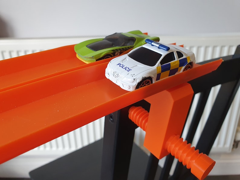 May include: Two toy cars on an orange track. The car in the front is a white police car with a blue light on top. The car in the back is a green sports car. The track is attached to a black and orange clamp.