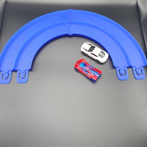 May include: Two toy cars on a blue plastic track. The track is curved and has a white car and a red and blue car on it.