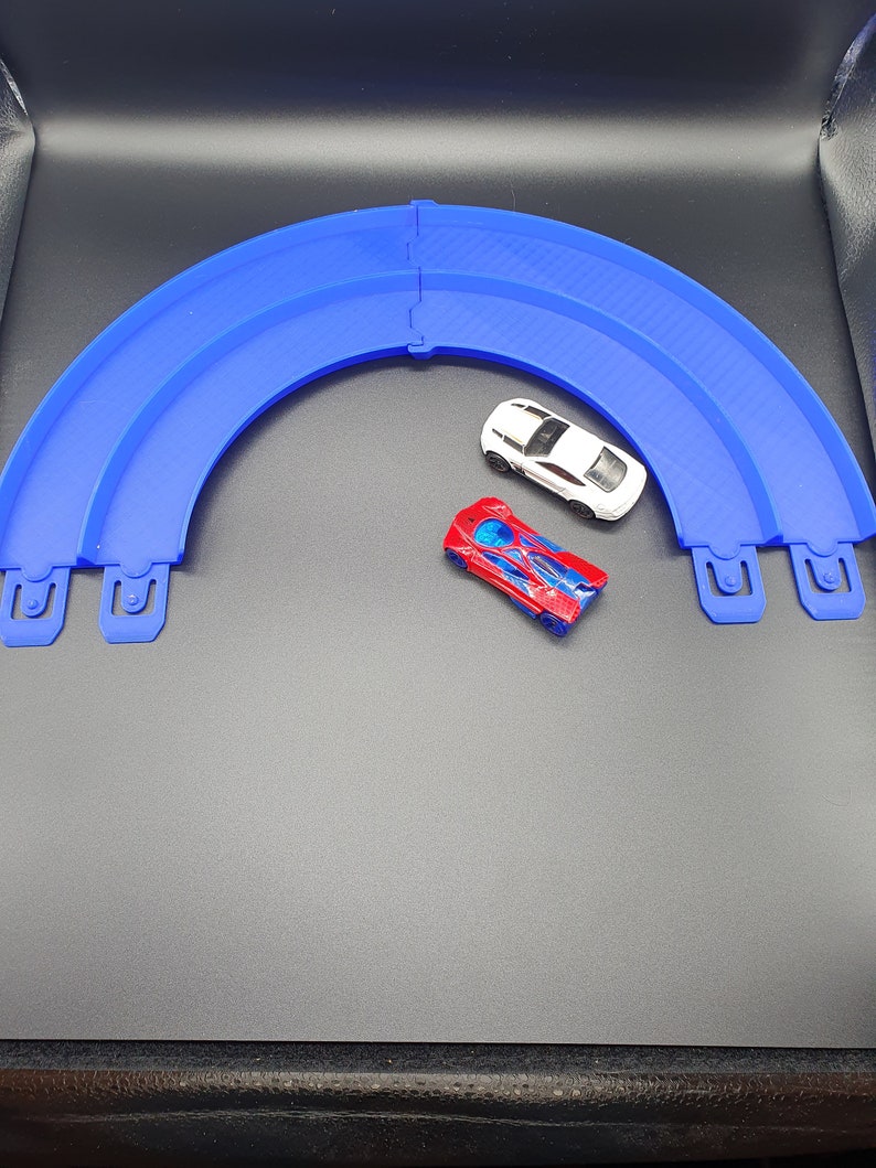 May include: Two toy cars on a blue plastic track. The track is curved and has a white car and a red and blue car on it.