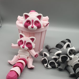 May include: Three 3D printed raccoon figurines. One is pink and sitting on a pink trash can. The other two are gray and lying down.