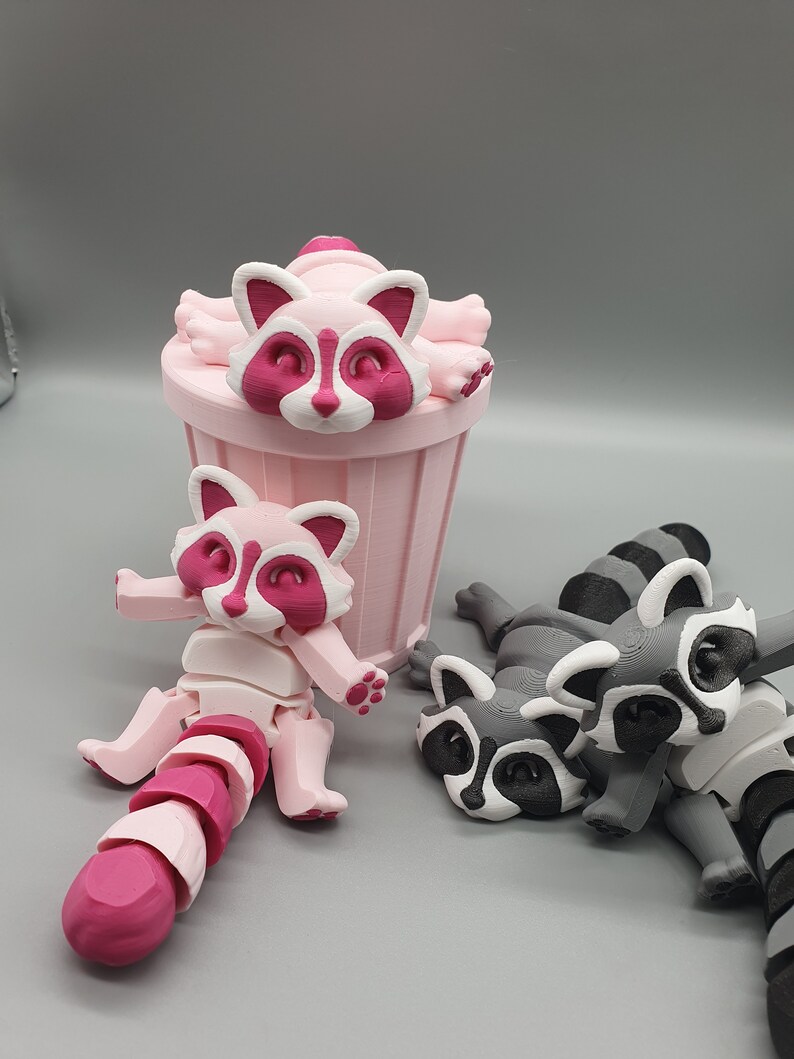 May include: Three 3D printed raccoon figurines. One is pink and sitting on a pink trash can. The other two are gray and lying down.