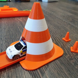 May include: A toy police car drives through a large orange and white traffic cone with a smaller orange traffic cone on either side.