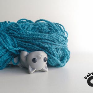 May include: A white plastic cat figurine with black eyes, sitting on a ball of teal yarn. The text "Baby CAT" is printed below the yarn.