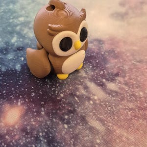 May include: A brown and white owl figurine with large black eyes. The owl is sitting on a purple and white galaxy-themed surface.